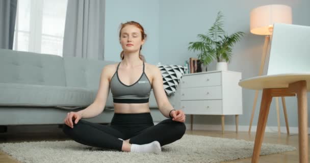 Young Caucasian woman with red hair sitting on floor in cozy living room and meditating. Pretty girl having meditation while doing yoga at home. Relax concept. - Felvétel, videó