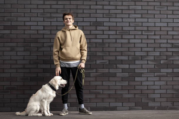 young guy with a puppy of a retriever stands on the street against the background of the wall and smiles, copy space - Zdjęcie, obraz