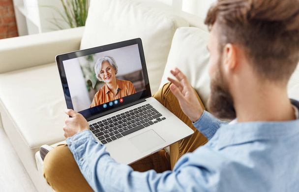 From above young male greeting elderly woman while sitting on couch and using video chat app on laptop at hom - 写真・画像