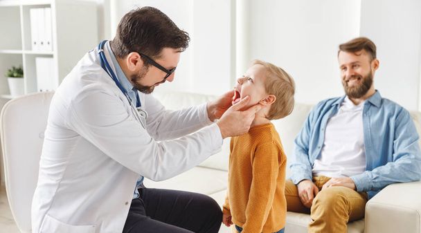Zijaanzicht van mannelijke arts inspecteren keel van kleine jongen in de buurt vader tijdens check-up afspraak in eigentijdse hospita - Foto, afbeelding