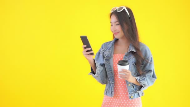 footage of beautiful Asian woman using smartphone and holding paper cup of coffee isolated on yellow - Záběry, video