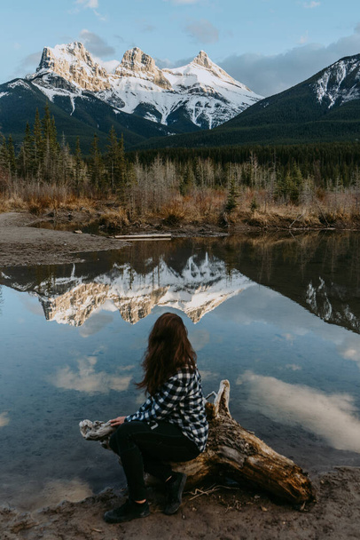 Toeristenvrouw kijkt naar het iconische de drie zusters bergtoppen reflecteren in Policeman 's Creek rustig water tijdens zonsondergang. Canmore, Alberta, Canada. - Foto, afbeelding