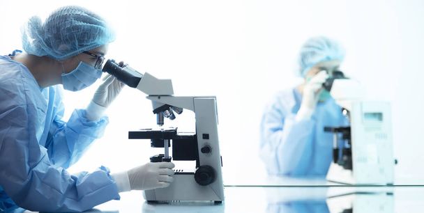 vrouwelijke artsen in pak voor bioveiligheid die virusanalyse doen onder een microscoop, pandemisch concept - Foto, afbeelding