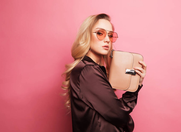 Young beautiful blond woman in a brown blouse holds a handbag and posing on pink background. - Photo, image