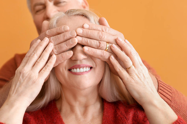 Happy elderly man covering eyes of his wife couple on color background, closeup - Zdjęcie, obraz