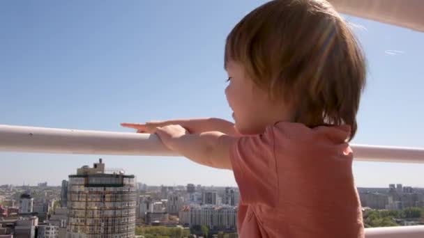 Mutlu anaokulu çocuğu bulutsuz mavi gökyüzünün altındaki gökdelenin balkonundan şehir manzarasına hayran. - Video, Çekim