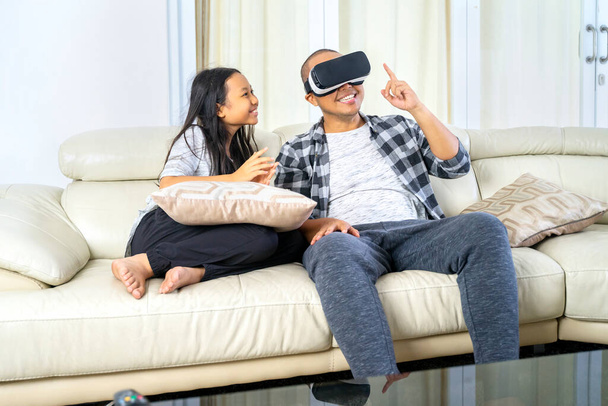 Young man wearing virtual reality headset to playing video games while sitting with his daughter on the sofa. Shot at home - Foto, Bild