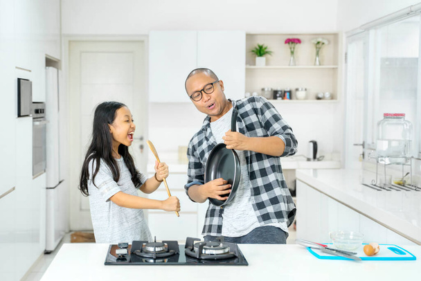 Young man with his daughter playing kitchenware like a microphone and guitar while having fun together in the kitchen at home. Shot in 4k resolution - Foto, Bild
