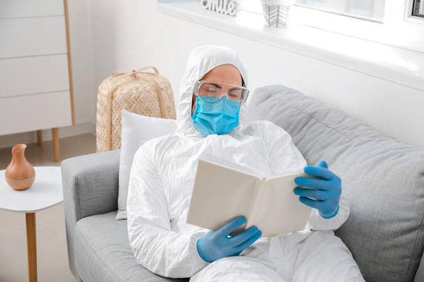 Biyolojik tehlike kıyafetli bir adam evde kitap okuyor. - Fotoğraf, Görsel