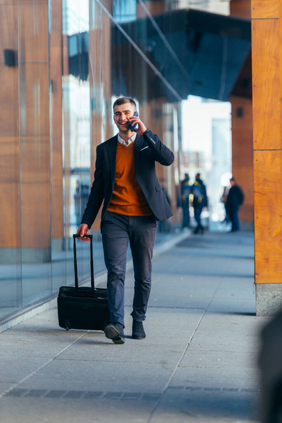 Nuori liikemies kävelee asemalla puhuessaan puhelimessa ja vetäessään matkalaukkuaan
. - Valokuva, kuva