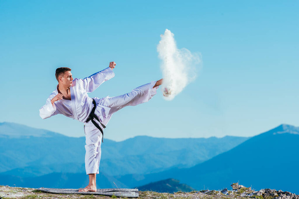Atleta de karate en forma pateando una taza llena de flúor causando un gran chapoteo mientras usa un kimono blanco en la cima de una montaña en un día soleado. - Foto, imagen