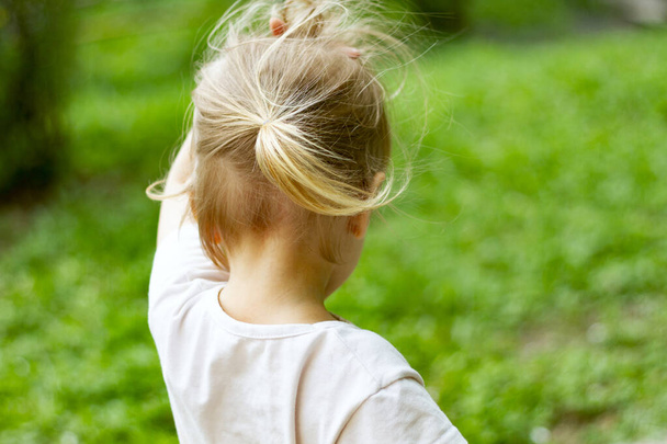 Adorable blond toddler girl in a windy summer day wearing ponytail trying to hold her fluterring hair. Veiw from behind. Copy space - Photo, image