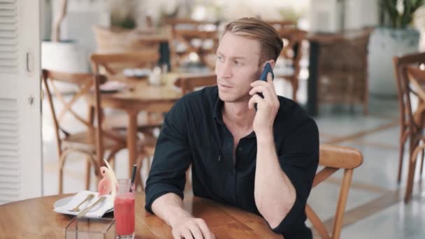 Portrait of businessman in black shirt sitting in cafe and talking on phone - Záběry, video