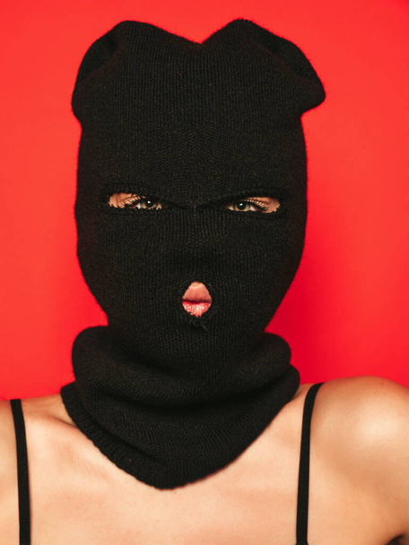 Beautiful sexy woman in black swimwear bathing suit. Model wearing bandit balaclava mask.Hot girl posing near red wall in studio.Seductive female in nice lingerie.Crime and violence - Photo, image