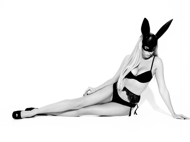 Portrait of young beautiful sexy woman with large breasts. Model wearing carnival black mask of Easter bunny rabbit.Hot blonde girl sitting in studio and isolated on white - Fotó, kép