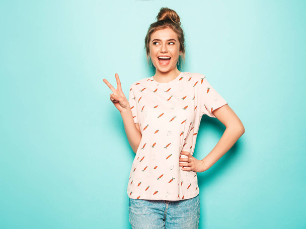 Young beautiful woman looking at camera. Trendy girl in casual summer T-shirt clothes showing peace sign. Positive female shows facial emotions. Funny model isolated on blue - Fotografie, Obrázek