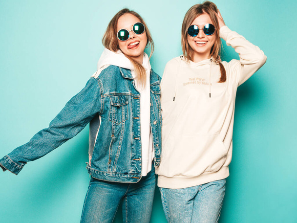 Two young beautiful brunette smiling hipster girls in trendy summer hoodie and jeans jacket clothes. Sexy carefree women posing near blue wall. Trendy and positive models having fun in sunglasses - Valokuva, kuva