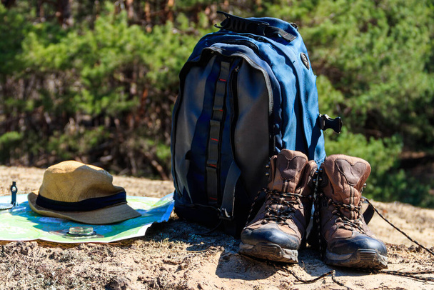 Plecak turystyczny z butami turystycznymi, kapeluszem, kompasem i mapą na polanie w sosnowym lesie. Urządzenia do wędrówek trekkingowych. Koncepcja podróży - Zdjęcie, obraz