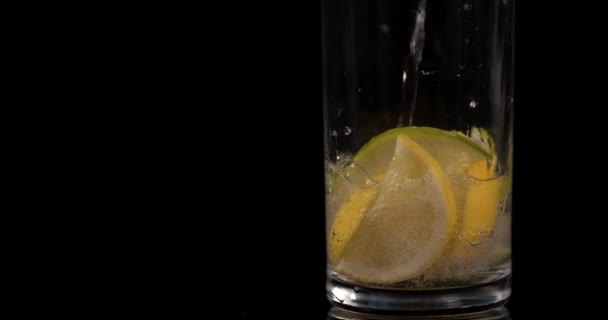 Close-up of pouring soda water with lemon and lime on a black background in slow motion. Lemonade with ice in glass - Footage, Video