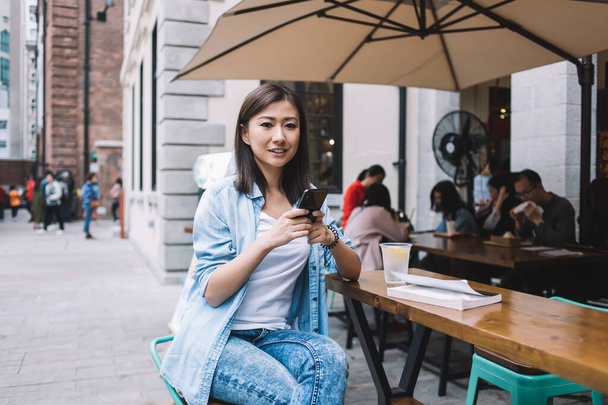 Happy brunette Asian woman in blue shirt texting on smartphone and looking at camera while sitting in outdoors cafe on wooden table with refreshing drink - Zdjęcie, obraz
