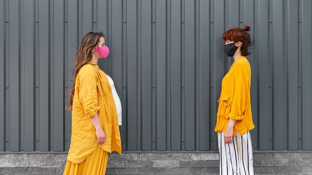 Dos mujeres con máscaras protectoras y ropa amarilla están de pie en la distancia. El concepto de distancia social. Fondo negro
 - Foto, Imagen