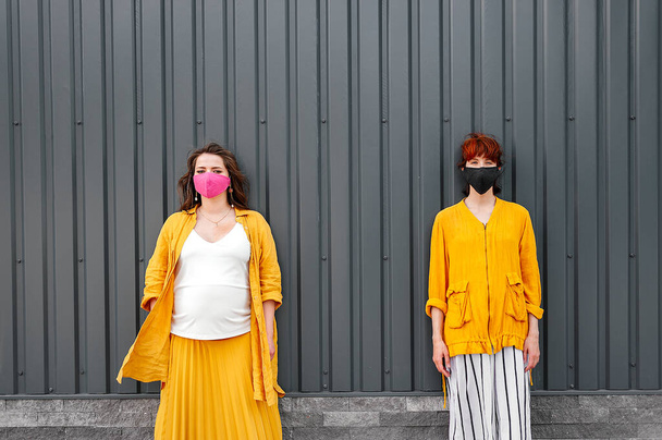 Δύο γυναίκες με προστατευτικές μάσκες και κίτρινα ρούχα στέκονται στο βάθος. Η έννοια της κοινωνικής απόστασης. Μαύρο φόντο - Φωτογραφία, εικόνα