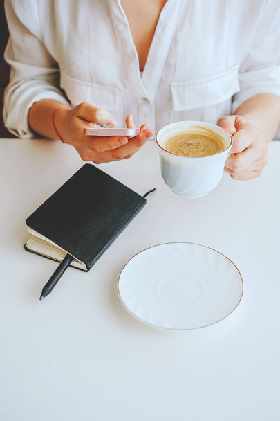 Το κορίτσι με το μαύρο μπουφάν κρατάει ένα τηλέφωνο και μια κούπα καφέ στο χέρι της. - Φωτογραφία, εικόνα