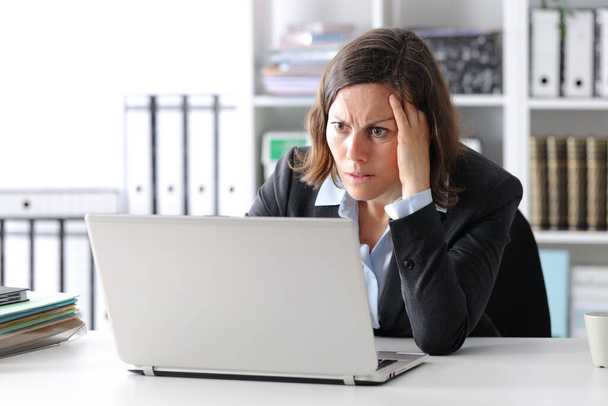 Worried adult executive woman reading news on laptop sitting on a desk at the office - Фото, изображение