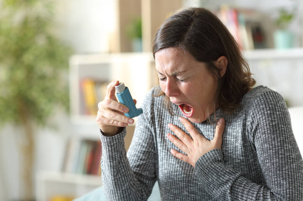 Asthmatic adult woman suffers asthma attack holding inhaler sitting on the couch at home - Foto, Bild
