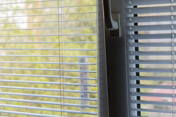 Фрагмент окна с горизонтальными жалюзи и прикрепленный внешний термометр снаружи, вид изнутри крупным планом в избирательном фокусе
  - Фото, изображение