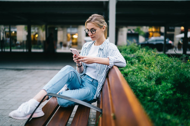 Vista lateral de la mujer rubia en ropa casual con anteojos que envían mensajes de texto en el teléfono inteligente mientras se relaja en el banco cerca de arbusto verde en el fondo borroso de la calle - Foto, Imagen