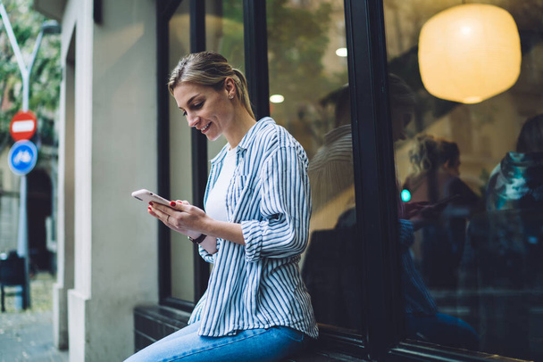 Side view of grinning blonde female in casual clothes sitting and browsing on smartphone near window outside restaurant on town street - Фото, изображение