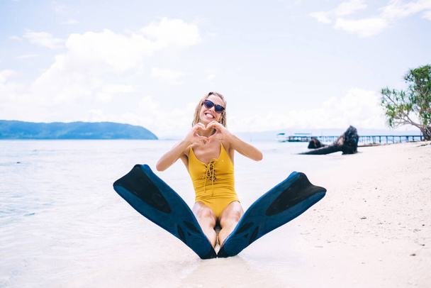 Young smiling blonde in yellow swimsuit, sunglasses and flippers sitting on beach and looking at camera against background of mountains, beach and palm trees while showing love gesture - Photo, Image