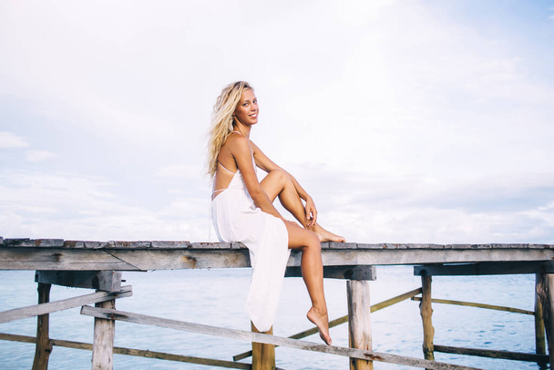 Delighted slim bronzed barefoot young blonde woman in white dress with open back sitting on high wooden pier at sea and looking at camera - Photo, Image