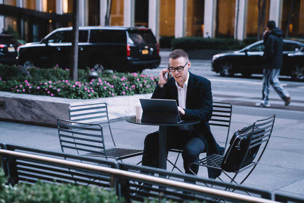 Male entrepreneur in formal attire with glasses talking on smartphone and working on tablet while sitting on downtown street with coffee - Foto, imagen