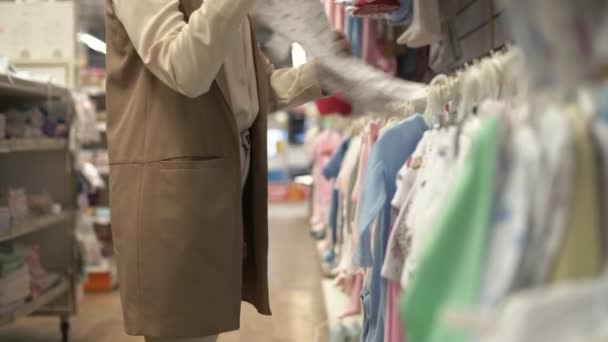 moederschap, jonge vrouwelijke klant in kinderkleding afdeling kiest dingen voor haar kind in de winkel, close-up - Video