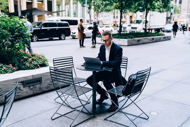 Presentable and focused executive in glasses and formal suit sitting outside in street cafe and browsing social media during break - 写真・画像