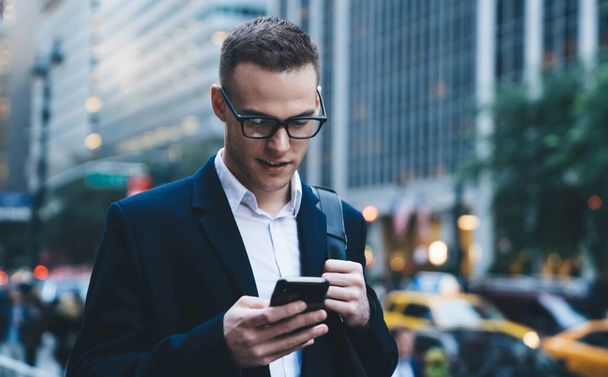 Confident young businessman in formal wear looking at phone and attentively reading information while walking down street and carrying bag - Photo, image