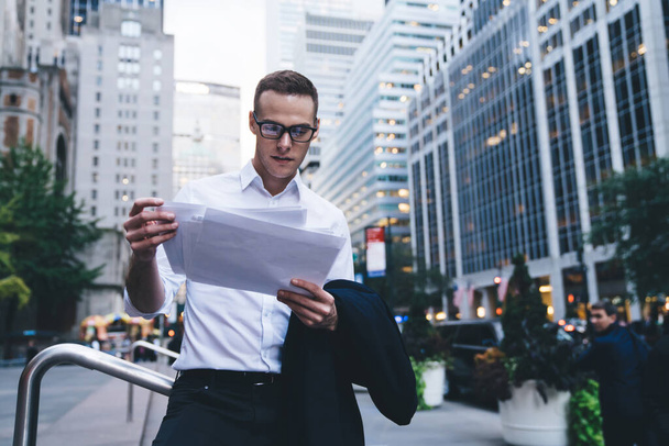 Young focused entrepreneur in formal wear and trendy glasses reading document while standing in middle of busy street in downtown on background of skyscrapers - Foto, immagini