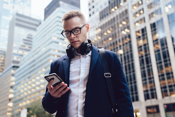 From below formal handsome executive in glasses and coat using wireless headphones and browsing phone on blurred background of commercial buildings  - Fotoğraf, Görsel