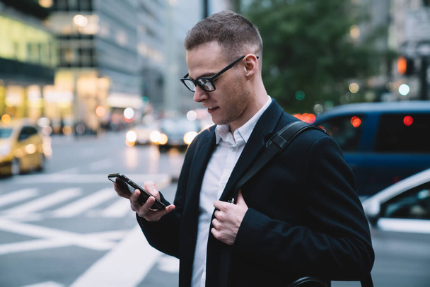 Merry and intelligent executive in glasses and formal suit texting message and smiling while standing in front of road on blurred background - 写真・画像