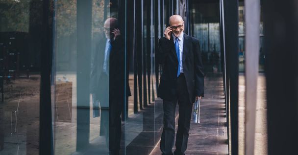 Smiling old businessman in formal outfit and eyeglasses and with white beard holding newspaper having phone call while walking in passage on street looking down - Photo, Image