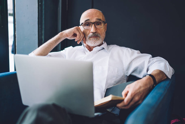 From above of elderly bearded bald man in white shirt and black pants with glasses working on laptop and making notes while looking at camera - Foto, Imagem