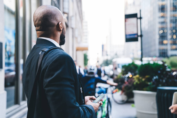 Back view of unrecognizable ethnic entrepreneur with black beard in classic suit standing at street with smartphone in hands and looking at city.Ethnic entrepreneur with smartphone in hands looking at city - Photo, image