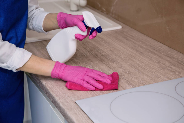 Ένα κορίτσι με ροζ λαστιχένια γάντια πλένει μια επιφάνεια του ντουλαπιού με ένα πανί μικροϊνών. - Φωτογραφία, εικόνα