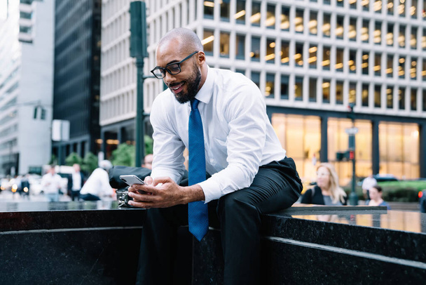 Cheerful formal bearded bald black businessman in white shirt and blue tie and with eyeglasses browsing mobile while sitting on street stairs on background of commercial buildings and people crowd - Φωτογραφία, εικόνα