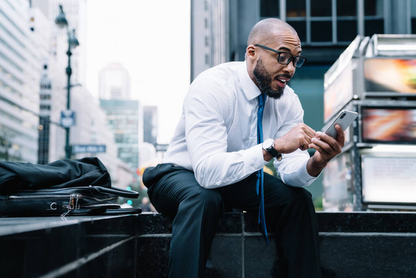 Heyecanlı, Afro-Amerikan erkek çalışanı telefonda şaşkınlıkla mesaj okurken arka planda deri bir çantayla, bulanık ticari binanın arka planında oturuyordu. - Fotoğraf, Görsel
