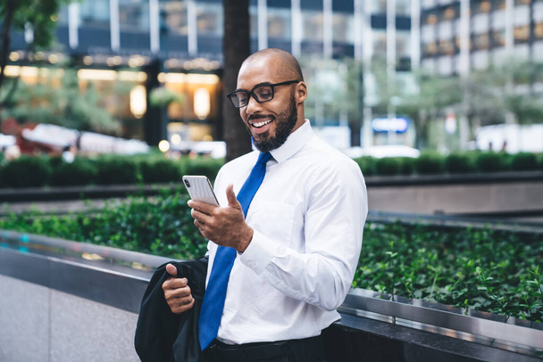 Homem étnico feliz em roupas de escritório e óculos segurando jaqueta terno e usando smartphone enquanto está em frente ao edifício de escritório moderno - Foto, Imagem