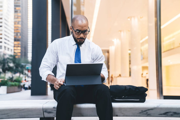 Occupied bald and bearded African American boss in white shirt with blue tie and eyeglasses sitting on stone bench and working on tablet - Foto, Imagem