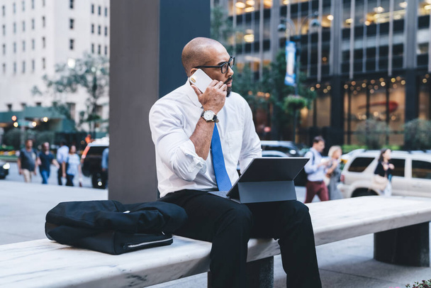 Bald African American male in formal wear and glasses sitting on white bench with jacket and leather bag holding tablet on knees and talking on mobile phone in downtown  - Foto, Imagem